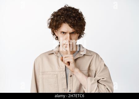 Primo piano di un uomo riccio arrabbiato che frena, rendendo il gesto di shush chiuso e fissando minaccioso sfondo bianco Foto Stock
