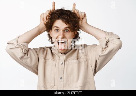 Ragazzo curly divertente e spensierato che mostra il gesto delle corna toro, bastone lingua e squinting, facendo volti, sfondo bianco Foto Stock