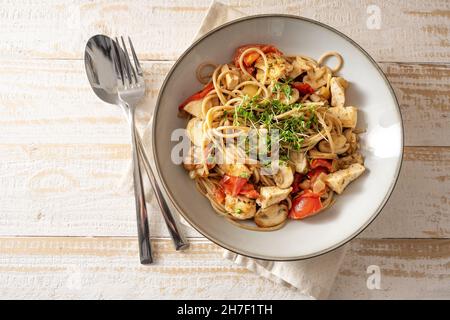 Spaghetti a grani interi con strisce di pomodoro, funghi e pollo, guarniti con cress, piatto grigio e posate su un tavolo di legno rustico chiaro, vista dall'alto Foto Stock