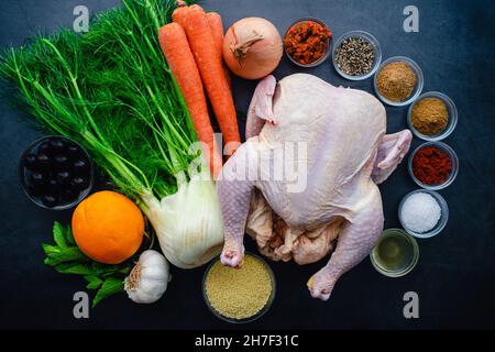 Ingredienti per il pollo speziato marocchino con verdure arrosto: Vista dall'alto di pollo crudo, verdure e spezie su sfondo scuro Foto Stock