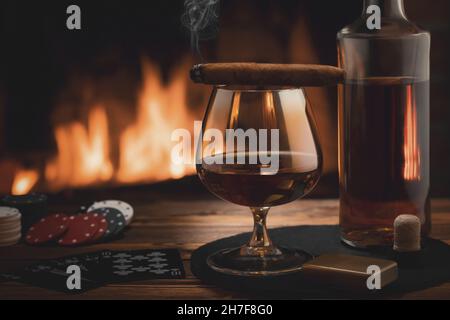 Bicchiere di cognac, sigaro, bottiglia, gioco di carte e fiches sul tavolo vicino al camino in fiamme. Riposo, piacere, buona fortuna e dolce vita concetto. Foto Stock