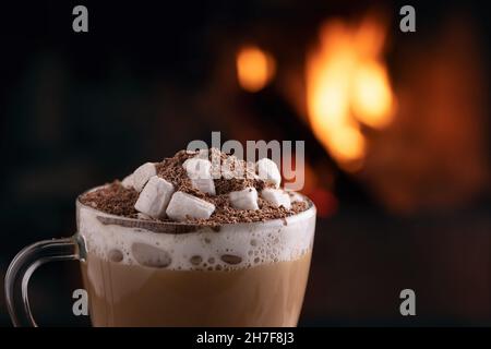 Cacao con marshmallows e cioccolato in una tazza di vetro su un tavolo di legno vicino ad un camino in fiamme. Foto Stock