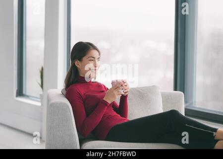 Home lifestyle Happy Asian donna rilassante bere tè in soggiorno godere di tempo libero nel fine settimana per rilassarsi alla finestra vista seduta in divano. All'interno Foto Stock
