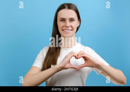 Felice giovane femmina fare con le dita cuore forma sorriso guardare la fotocamera attraverso il simbolo, in posa isolato su sfondo blu Foto Stock