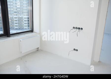 Una moderna ristrutturazione delle camere con pareti bianche e radiatore e una cornice nera delle finestre. Installazione incompleta di prese elettriche, spine e interruttori A. Foto Stock