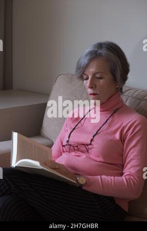 Donna anziana in occhiali appoggiati sul divano e leggendo un libro. Concetto hobby. Foto Stock