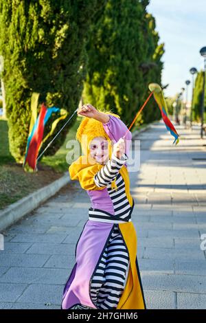 Felice clown femminile con volto dipinto in costume divertente e parrucca gialla, sorridente e camminando sul sentiero nel parco con poi colorato nelle mani durante la sfilata Foto Stock