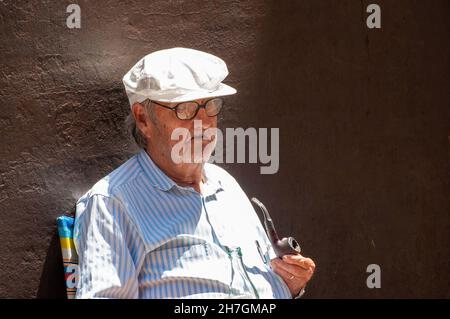 Spagna-Luglio 2020: Primo piano di un uomo più anziano camicia a strisce blu e cappuccio bianco seduta in una sedia solare colorata contro una parete scura che fuma un tubo Foto Stock