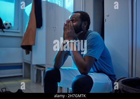 Teso giovane giocatore di calcio in uniforme sportiva mantenendo le mani unite dal viso mentre si siede da armadietti in spogliatoio Foto Stock