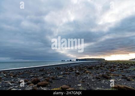 Vista panoramica sulla spiaggia vulcanica di sabbia nera di Reynisfjara in Islanda con alcune formazioni rocciose rimaste nell'oceano e le nuvole scure nel cielo durin Foto Stock