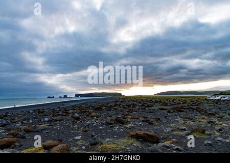 Vista panoramica sulla spiaggia vulcanica di sabbia nera di Reynisfjara in Islanda con alcune formazioni rocciose rimaste nell'oceano e le nuvole scure nel cielo durin Foto Stock
