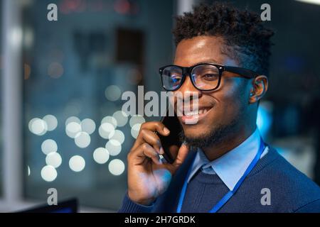 Felice giovane operaio africano in occhiali e maglione blu che parla da telefono cellulare in grande ufficio open space Foto Stock