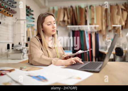 La designer femminile utilizza il laptop sulla sua scrivania, che è coperto da vari schizzi. Vestiti appendere e schizzi pinned al muro del suo studio o a. Foto Stock