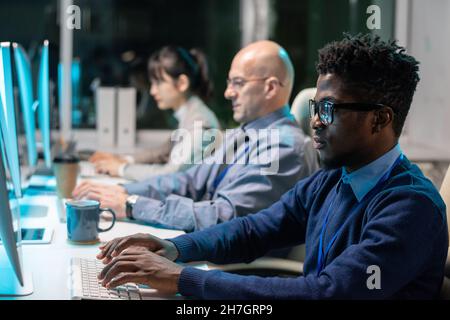 Fila di programmatori interculturali contemporanei che lavorano su nuovi software di fronte a monitor di computer in ufficio Foto Stock