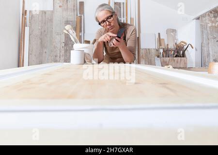 artigianato del legno, falegname artigianale donna con telefono cellulare cerca in internet nel suo laboratorio, restauro, fai da te e opere fatte a mano concetto Foto Stock