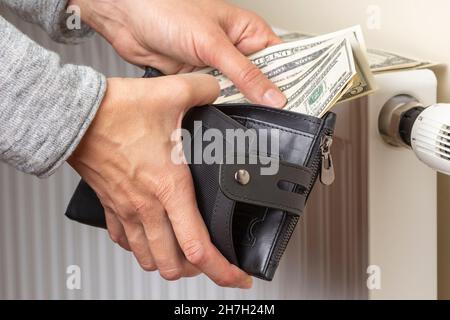 La mano della donna prende le banconote in dollari statunitensi dal portafoglio e mette sulla batteria del radiatore di riscaldamento con regolatore di temperatura. Costi di riscaldamento costosi durante Foto Stock