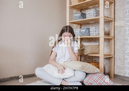 una giovane ragazza che piange copre il viso con le mani mentre si siede sul pavimento in una stanza tra cuscini. tristezza e tristezza Foto Stock