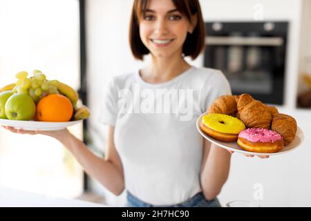 Dieta sana vs cibo spazzatura. Graziosa signora che tiene frutta e ciambelle, tentata di mangiare dessert invece di frutta Foto Stock