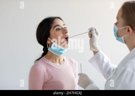 Medico europeo che fa il test della pcr covid-19 per la giovane donna indiana su sfondo grigio della parete in clinica Foto Stock