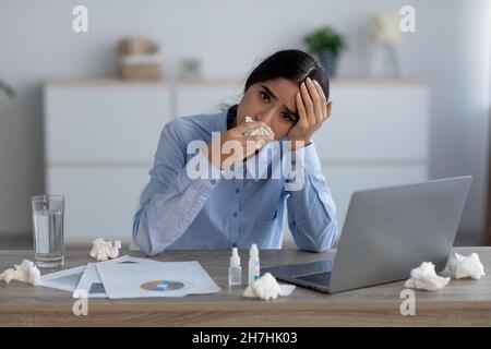 Stanca infelice triste giovane donna indiana che soffre di malattie e strofinando il naso con tovagliolo sul posto di lavoro Foto Stock