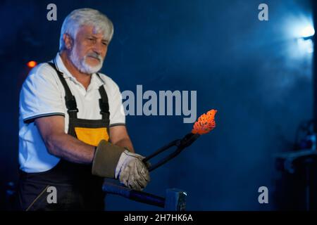 Fabbro caucasico qualificato che forma l'acciaio fuso con utensili industriali a fucina. Uomo dai capelli grigi in grembiule protettivo e guanti forgiando manualmente il metallo. Foto Stock