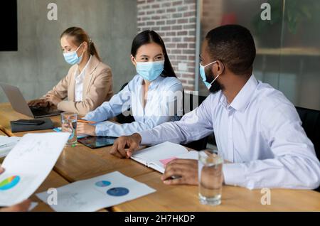 Colleghi multietnici che indossano la maschera medica che comunicano durante la riunione in ufficio Foto Stock