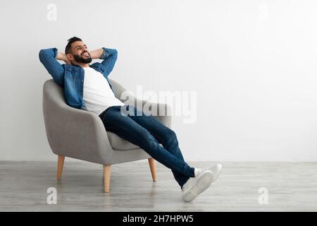 Calma giovane uomo arabo rilassarsi in poltrona contro il muro bianco studio, spazio copia Foto Stock
