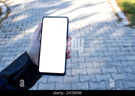 Immagine mockup di una mano donna che tiene il telefono cellulare con schermo bianco vuoto mentre si cammina sul marciapiede. Foto Stock