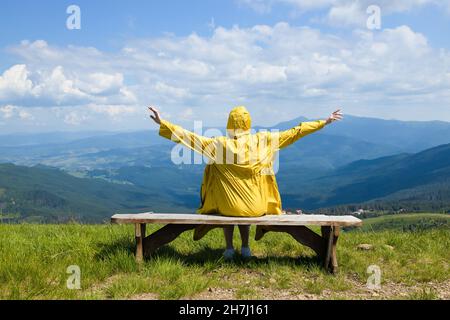 L'escursionista femminile di avventura alza le mani si siede su una panchina e gode di vista panoramica di montagna in una giornata estiva soleggiata Foto Stock