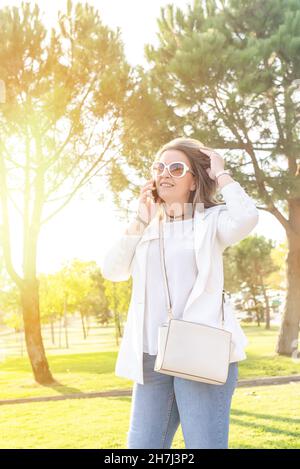 Donna caucasica di mezza età in piedi in un parco sorridendo e parlando sul suo telefono cellulare mentre le tocca i capelli Foto Stock