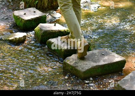 primo piano delle gambe e dei piedi dell'uomo che camminano su pietre a forma di rettangolo che si stagliano su un ruscello nel bosco. immagine della campagna al sole e all'ombra Foto Stock