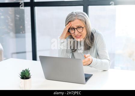 Donna anziana dai capelli grigi turbati che guarda lo schermo del laptop seduto alla scrivania in ufficio, che legge la notifica delle cattive notizie, donna d'affari matura frustrato ha commesso un errore nel lavoro, ha problemi con il progetto Foto Stock