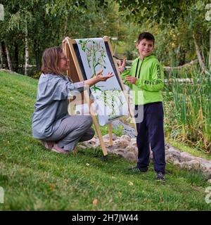 La madre insegna a ragazzo figlio di dipingere su tela. Donna insegnante artista dipinge con un bambino su carta natura e alberi presso il fiume Foto Stock