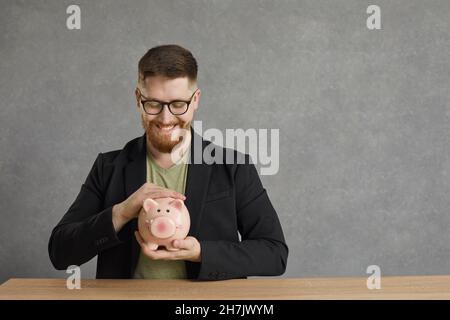 Sorridente giovane uomo che tiene la banca di maiale piggy, uomo seduto al tavolo Foto Stock