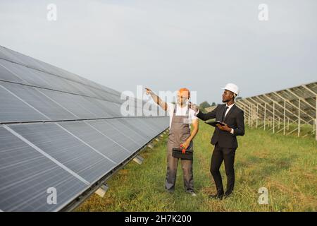 Uomo afro-americano in abito nero e casco bianco a piedi con tecnico indiano in uniforme su fattoria solare. Persone multirazziali che esaminano la produzione di energia verde pulita. Foto Stock