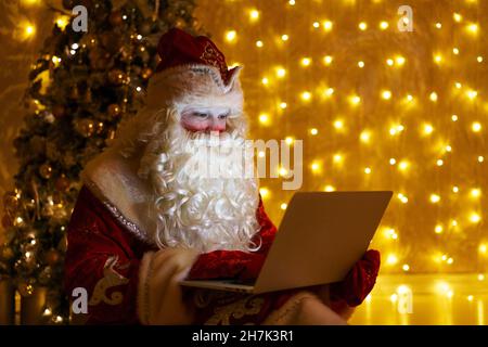 Auguri online di Natale da babbo natale. Babbo natale usando il portatile in distanza natale chiamare i bambini seduti vicino albero di natale a casa. Felice anno nuovo covid. Sullo sfondo di muro con ghirlanda Foto Stock