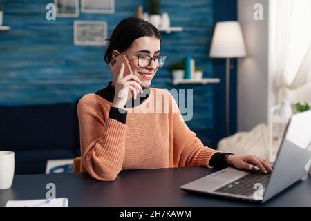 Sorridendo lo studente con gli occhiali utilizzando lo smartphone e navigare sul laptop. Freelance in ufficio a casa che parla al telefono mentre interagisce con il computer. Programmatore che utilizza la tecnologia per il lavoro remoto. Foto Stock