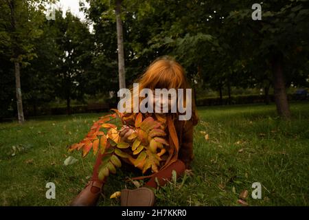 Sorridente ragazza rossa con le foglie che siedono sull'erba al parco Foto Stock