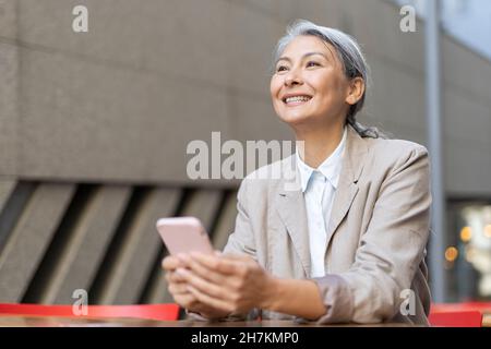 Donna matura sorridente che tiene il telefono cellulare al caffè sul marciapiede Foto Stock