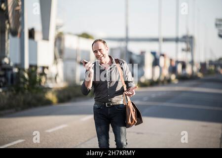Un uomo d'affari felice che invia messaggi vocali tramite smartphone mentre cammina per strada Foto Stock