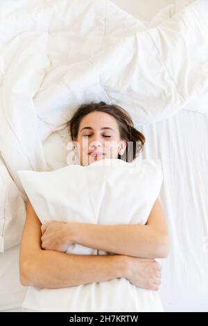 Sorridente giovane donna che abbraccia il cuscino mentre dorme a letto Foto Stock