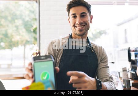 Cameriere sorridente che mostra il simbolo del segno di spunta sullo schermo del lettore di schede al bar Foto Stock