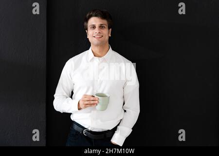 Giovane uomo d'affari con una tazza di caffè appoggiata sulla parete nera in ufficio creativo Foto Stock