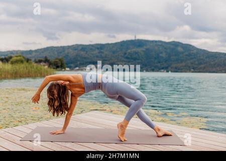 Donna attiva che pratica yoga sul molo vicino al lago Foto Stock