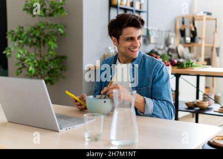 Giovane uomo seduto con un computer portatile che mangia tagliatelle a tavola Foto Stock