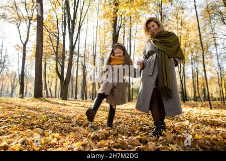 Donna sorridente che tiene la mano della figlia calci le foglie d'autunno nel parco Foto Stock