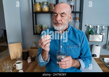 Uomo che tiene un bicchiere d'acqua prendendo la medicina dalla scatola della pillola a casa Foto Stock