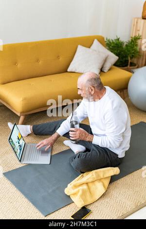 Uomo sorridente con computer portatile e bottiglia d'acqua seduti a casa Foto Stock