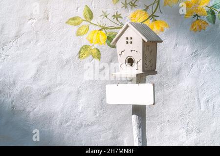 Bella scatola di nidificazione in legno con piastra bianca bianca bianca bianca appesa, sfondo di parete bianca testurizzata con fiore dipinto, spazio vuoto per il testo, raggio di luce an Foto Stock