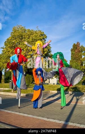 Gruppo completo di clown felici con volti dipinti in costumi colorati che tengono le mani e si divertono mentre si alza su palafitte nel parco, in giornata di sole Foto Stock